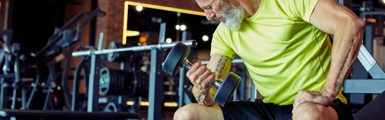 Gaining muscles after 40. Strong mature man in sportswear lifting heavy dumbbells and pumping his...