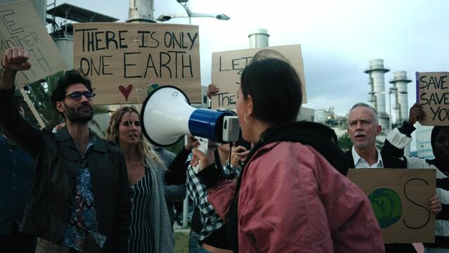 Rebel young activist woman speaking at a protest holding a megaphone to the group of people at a demonstration against pollution and factories with several banners of climate change messages