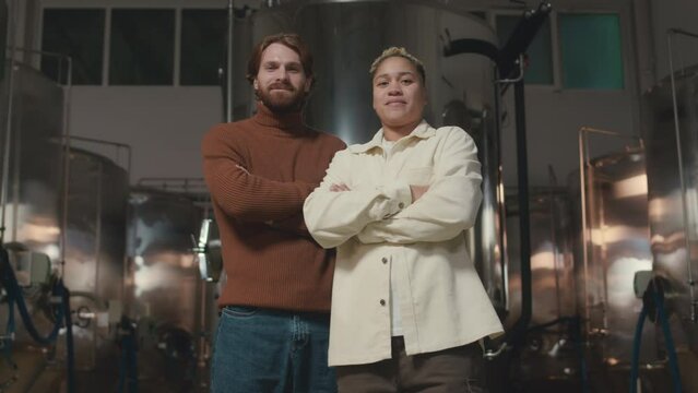 Low angle medium portrait of two confident ethnically diverse male and female beer technicians posing for camera with hands folded at brewing factory