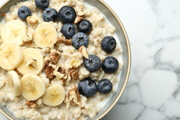 Tasty oatmeal with banana, blueberries, walnuts and milk served in bowl on white marble table, top view