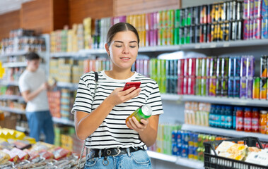 Casually dressed young brunette making purchases in grocery store, using smartphone to scan QR code on tin can of soft drink to get information about product. Modern shopping concept