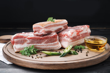 Pieces of raw pork belly, chili pepper, peppercorns, oil and parsley on black wooden table, closeup