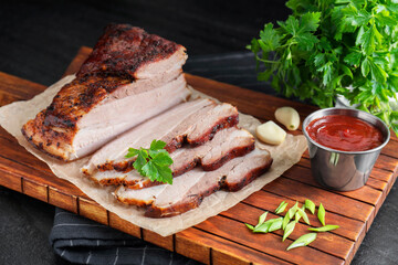 Pieces of baked pork belly served with sauce and parsley on black textured table, closeup