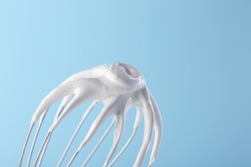 Whisk with whipped cream on light blue background, closeup. Space for text