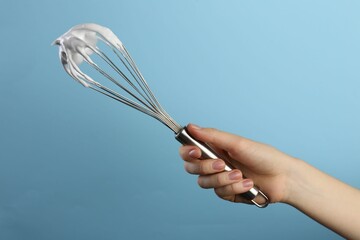 Woman holding whisk with whipped cream on light blue background, closeup
