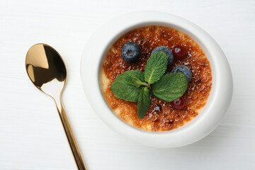 Delicious creme brulee with berries and mint in bowl and spoon on white wooden table, top view
