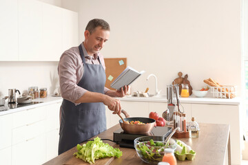 Mature man frying vegetables with recipe book in kitchen - obrazy, fototapety, plakaty