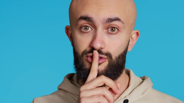 Middle eastern person showing mute gesture with finger over lips, posing over blue background. Arab guy doing hush symbol to keep silence, trying to share a secret whispering. Camera 2.