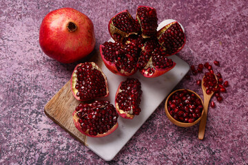 Board with fresh pomegranates and bowl of seeds on purple background