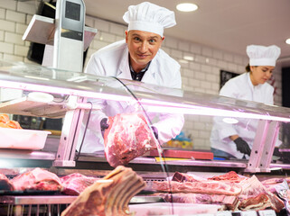 Smiling skilled adult butcher working behind counter in butchery, offering piece of fresh raw beef...