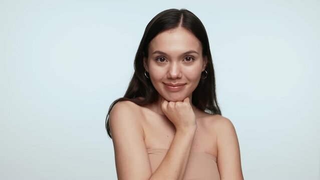 A fresh-faced young woman in her 20s poses gracefully against a light blue background, embodying beauty and youth with her clear skin, gentle smile, and natural elegance.