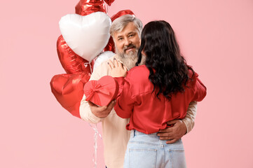 Beautiful mature couple with heart-shaped balloons and gift box hugging on pink background....