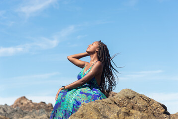 Beautiful woman in blue clothes sitting on the rocks on the beach sunbathing and touching her braids