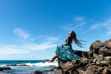 Beautiful woman in blue clothes and braided hair sitting on the edge of the beach rocks sunbathing