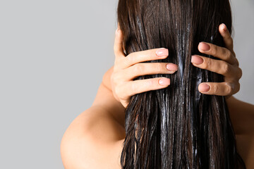 Young woman using cosmetics for hair treatment on grey background