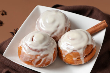 Plate with glazed buns, napkin and cinnamon sticks on brown background