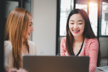 Two joyful businesswomen sharing a pleasant work moment on a laptop in a bright, collaborative office environment.