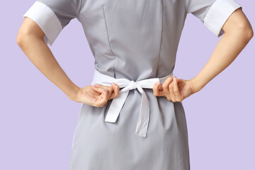 Young chambermaid tying apron on lilac background, back view