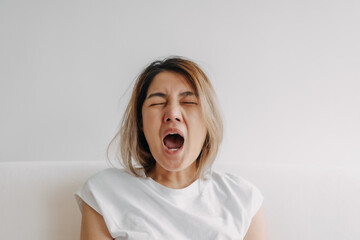 Sleepy face Asian woman yawning and rubbing her eyes in white t-shirt sits on the sofa.