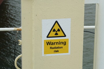 Label Warning radiation risk in shape of yellow triangle with black symbol of radiation is placed to electric box of big antenna situated on the ship. In yellow field is written with black letters. 