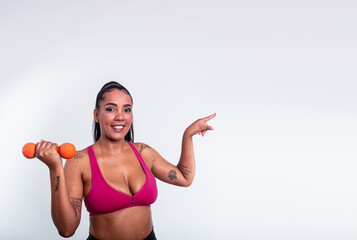 Beautiful woman dressed in gym clothes holding dumbbells and pointing finger to the side.