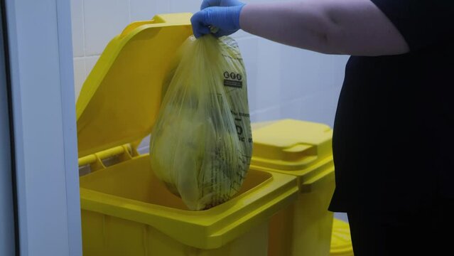 4K video. The concept of medical waste, a female hand in blue gloves opens a yellow tank and lowers a garbage bag. Disposal and storage of hospital waste