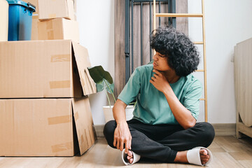Asian man sitting cross legged on floor and looking at stack of cardboard boxes in new apartment 