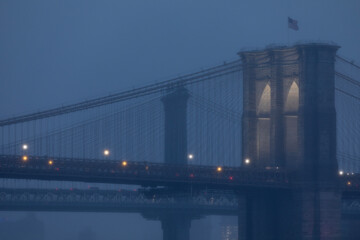 Foggy evening view of the Brooklyn Bridge illuminated at dusk.