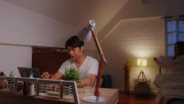Young asian man overworking at home using laptop computer at night in bedroom. His Asian girlfriend in pajamas stands and gives him a massage, encouraging him to finish his hastily completed work.