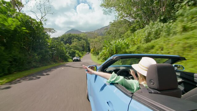 Woman driving in botanical tropical garden on Hawaii road trip. Free girl with hand out of convertible car rides with wind blowing in face. Stylish tourist stretches hand out of rent car without roof