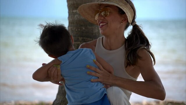 Slow motion of a latin mother woman playing with her son sitting next to a palm tree having fun and smiling