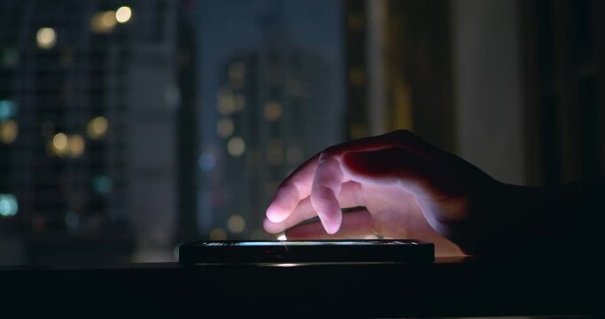 Close-up of person hand interacting with mobile phone lying on table. Late in evening, man surfs social networks on phone near window overlooking city. Glowing mobile phone screen in twilight.
