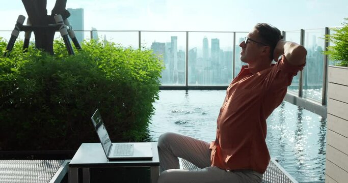 Man stretches sitting laptop near pool against cityscape. Remote work, blending relaxation productivity, embodies new era remote work. captures comfort flexibility remote. urban setting