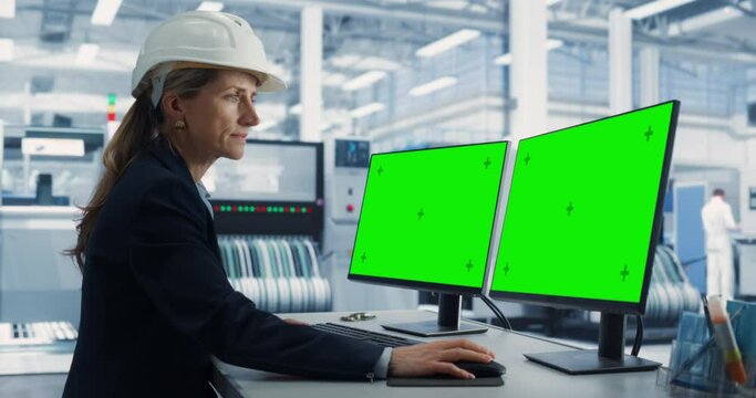 Caucasian Female Technician In Hardhat Working on a Desktop Computer with Green Screen Chromakey Mock-Up Display in a Factory Office. Autonomous Technological Research and Development Facility.