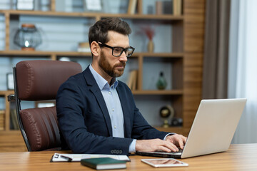 Focused male in eyeglasses doing computer work while sitting in leather chair by wooden desk in...