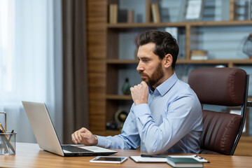 Focused bearded man looking attentively at portable computer screen and holding hand under chin in...