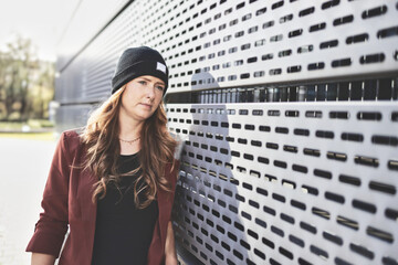 Woman in her mid-thirties with long red hair wearing black clothing with a brown jacket and hat in an urban environment.