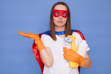 Upset disappointed brown haired woman wearing superhero costume mask and orange rubber gloves posing isolated over blue background holding sanitary supplies spreading palm copy space for advertisement © sementsova321