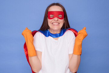 Overjoyed Caucasian brown haired woman wearing superhero costume white T-shirt and rubber gloves clenched fists rejoicing her success isolated over blue background