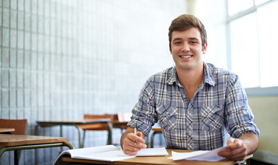 Student, man and portrait in classroom for education, learning and writing of knowledge in university. Face of young and smart person with notes for college research, essay paper and studying at desk - obrazy, fototapety, plakaty