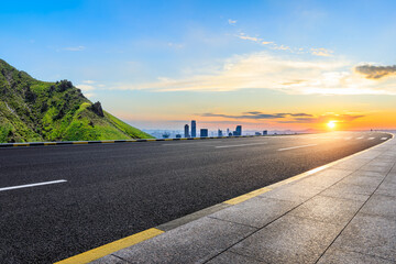 Asphalt highway road and green mountain with city skyline at sunset - obrazy, fototapety, plakaty