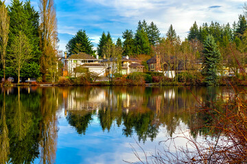 Lake Como Park in Coquitlam on a sunny winter day. A village on the shore of a lake surrounded by coniferous trees. Reflection of houses and green trees in mirror water against a blue cloudy sky