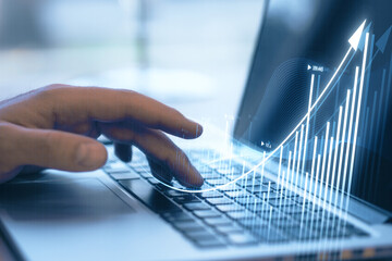 Close up of businessman hand using laptop keyboard with growing business chart hologram on blurry...