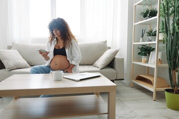 Pregnant woman blogger sits on the couch at home and takes pictures of herself on the phone, selfie and video call, consultation with the doctor online, pregnancy management, freelance work