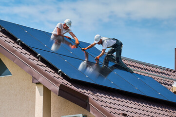 Workers building solar panel system on rooftop of house. Two men installers in helmets installing...