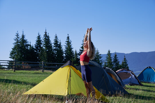 Woman tourist camping outdoors. Slim, sporty lady standing near tent with closed eyes, stretching, raising hands, smiling, wearing sportswear. Concept of tourism and hiking in mountains.