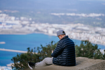 Gibraltar, Britain - January 24, 2024 - A man in a blue jacket and cap sits looking at a coastal city from a high vantage point.