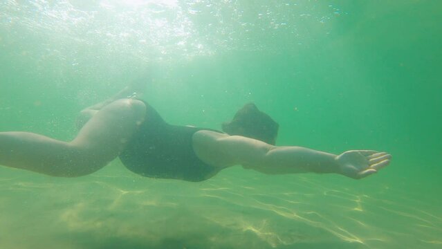 Pregnant, beautiful woman in a black swimsuit swims underwater in the tropical sea, in which the rays of the sun play. Healthy and active pregnancy concept. Water activities, travel during pregnancy