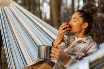 A beautiful curly-haired Caucasian hiker unwinds in her hammock, relishing a coffee and apple as a hiking snack.