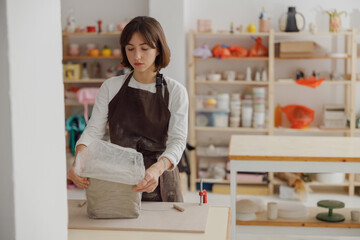 Young woman preparing clay to create a mug on a wooden table in pottery studio